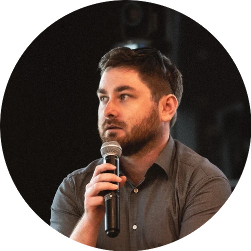 Callum holding a microphone at a speaking event, wearing a dark shirt with sunglasses resting on his head.